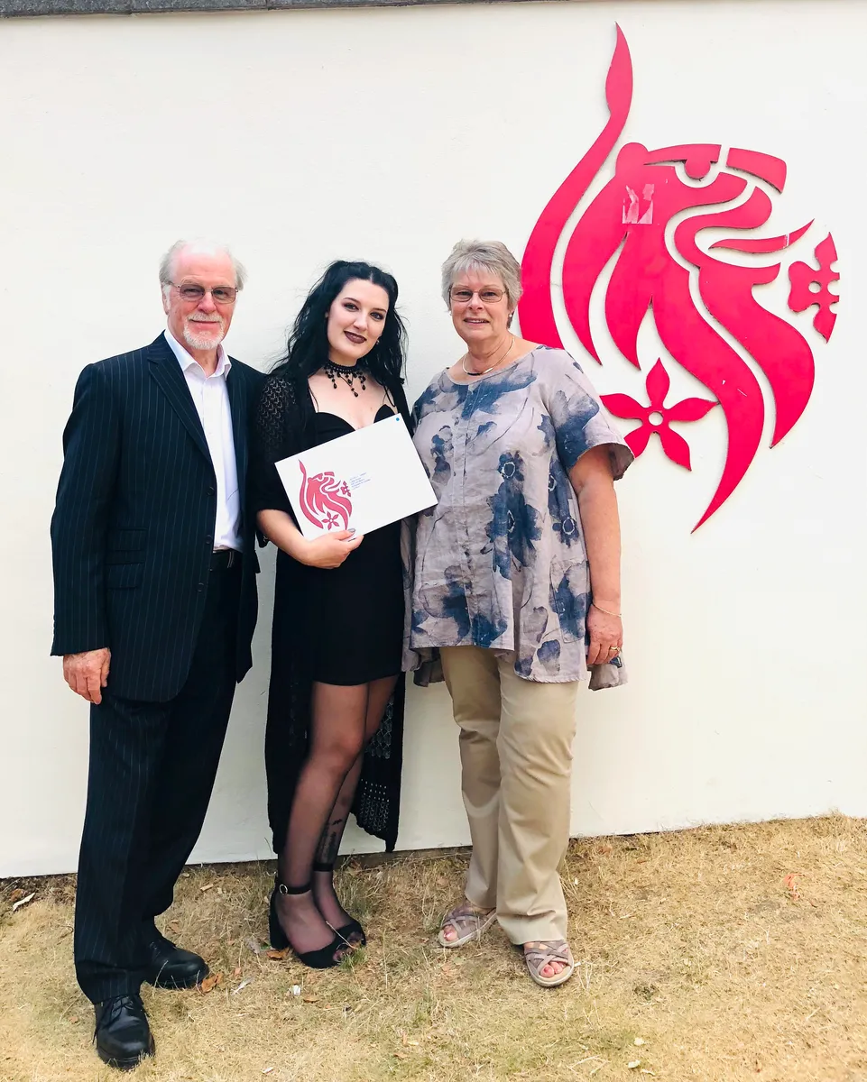 Cassandra at her graduation, holding her degree certificate, with family either side